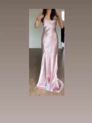 Lång rosa satinklänning - Elegant långklänning i skimrande rosa satin med smala axelband och figurnära passform. Klänningen har en mjukt draperad urringning och faller snyggt längs kroppen för en lyxig känsla. ryggen är öppen längre upp med bara banden till armarna samt en knytning. En dragkedja finns på baksidan men syns inte stängd. Köptes i mars förra året och användes en gång till min bal i juli. Släp på klänningen finns som syns någorlunda bra på bild 3. Även en slits på sidan av klänningen.