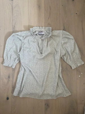 Prickig topp - Supersöt vit blus från Holly & Whyte med små svarta prickar, volangkrage och knäppning vid halsen. Blusen har trekvartsärm med lätt puff och smock vid ärmslut. Tillverkad i ett tunt, mjukt tyg som känns luftigt och skönt. Storlek: S❣️