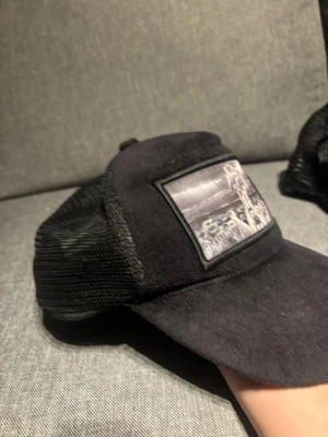 Svart truckerkeps från Berretto Sweden - Snygg svart truckerkeps från Berretto Sweden med mesh på sidorna och bak, samt ett svartvitt tryck framtill. Justerbar baktill för perfekt passform. Kepsen har en böjd skärm och är tillverkad i en mix av tyg och meshmaterial.