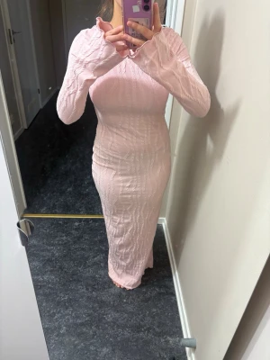 Ljusrosa långklänning med öppen rygg - Supersnygg ljusrosa långklänning med stickad struktur och lång ärm. Klänningen har en öppen rygg med knytband upptill som ger en extra detalj. Perfekt för dig som vill ha något unikt och trendigt.