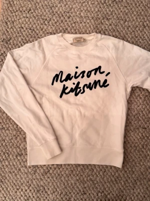 Vit sweatshirt Maison Kitsuné🙌 - Vit sweatshirt från Maison Kitsuné med svart handskriven logga på bröstet. Tröjan har rund halsringning, långa ärmar och ribbade muddar vid ärmslut och nederkant. Tillverkad i mjukt bomullsmaterial för en skön och avslappnad look.🙌