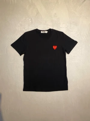 Svart Comme des Garçons PLAY t-shirt - Säljer en svart t-shirt från Comme des Garçons PLAY med det ikoniska röda hjärtat med ögon broderat på bröstet. T-shirten har rund hals och korta ärmar. Tillverkad i mjuk bomull som känns skön mot huden. Perfekt för dig som gillar stilrena plagg med en twist.