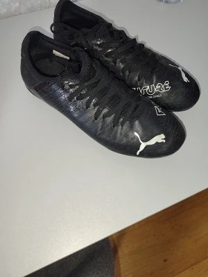 Svarta Puma Future fotbollsskor - Säljer ett par svarta Puma Future fotbollsskor med vita detaljer och logga. Skorna har snörning och en smidig, låg profil som ger bra bollkontroll. Tillverkade i syntetmaterial för lätt vikt och bra passform. Perfekta för dig som vill ha stilrena och funktionella skor på planen. Dubbade . 