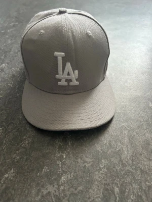 Grå LA-keps från New Era - Snygg grå keps från New Era med LA Dodgers-logga broderad framtill i vitt. Klassisk flat brim och broderad detalj på sidan. Perfekt för dig som gillar streetstyle och sportiga accessoarer.