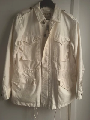 Vit utilityjacka från Polo Ralph Lauren - Stilren vit utilityjacka från Polo Ralph Lauren med fyra stora fickor framtill, dragsko i midjan och nederkant samt axelklaffar. Jackan har klassisk krage och knäppning med både dragkedja och knappar. Perfekt för en avslappnad men snygg look. 