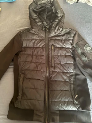 Svart pufferjacka Supply & Demand - Svart pufferjacka med huva från Supply & Demand. Jackan har quiltad framsida, släta ärmar, dragkedja framtill och en bröstficka med dragkedja. På vänster ärm sitter ett runt tygmärke med logga. Perfekt för kalla dagar. Den är pyttelite solbränd