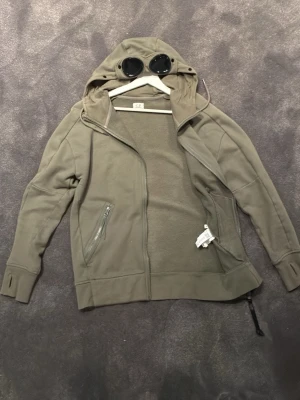 Olivgrön hoodie med goggles från C.P. Company - Säljer en olivgrön hoodie från C.P. Company med dragkedja och ikoniska goggles i huvan. Hoodien har två fickor med dragkedja och är tillverkad i mjukt bomullsmaterial. Perfekt för dig som gillar streetwear och unika detaljer. Hör av er vid frågor! 👍