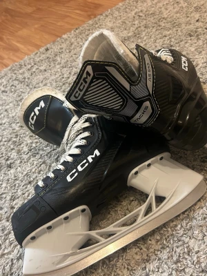 CCM AS 555 hockeyskridskor svart/vit - Säljer ett par svarta och vita CCM AS 555 hockeyskridskor med vita snören och robust design. Skridskorna har en modern look med detaljer i silver och svart samt en stabil plastkonstruktion runt bladet. Perfekta för dig som gillar att åka skridskor på isen.