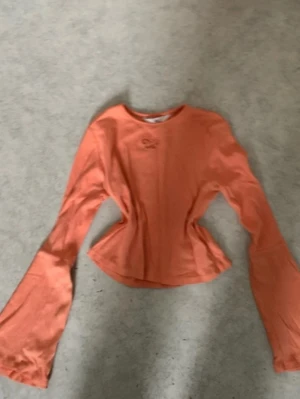 Orange långärmad topp  - Trendig orange topp med långa utsvängda ärmar och en söt  vid halsen. Figurnära passform som framhäver midjan. Perfekt för dig som vill sticka ut med färg och form.