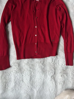 Röd kofta från Lindex  - Snygg röd cardigan från Lindex med långa ärmar och knappar framtill. Tillverkad i ett mjukt, tunt material som passar perfekt till lager på lager. Klassisk passform och ribbade muddar vid ärmslut och nederkant.