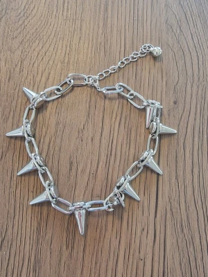 Kedjehalsband med silverfärgade spikes - Säljer ett chunky halsband i silverfärgad metall med stora länkar och flera spetsiga spikes runt om. Halsbandet har justerbar kedja och en liten pärldetalj vid låset. Perfekt för en edgy stil.Halsband punk. Längd 35 cm. Spikar 2 cm.