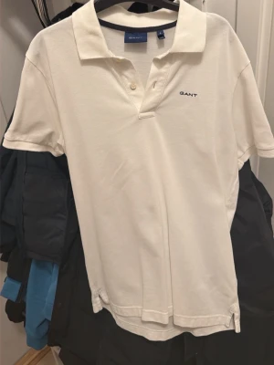 Vit pikétröja från GANT - Klassisk vit pikétröja från GANT med korta ärmar och krage. Tröjan har två knappar vid halsen och diskret GANT-logga broderad på bröstet. Perfekt till jeans eller shorts för en clean look.