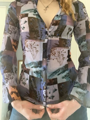 Mönstrad meshskjorta med botaniskt tryck - Säljer en långärmad meshskjorta med coolt botaniskt patchwork-mönster i lila, brunt, blått och grönt. Skjortan har genomskinligt material, knappar framtill och figurnära passform . Står storlek S, men är mer XS. Är helt oanvänd och passar mindre bröst.