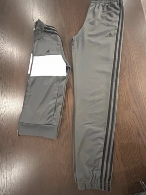 Grå Adidas träningsset med vita detaljer - Säljer ett grått träningsset från Adidas med klassiska svarta ränder längs ärmar och ben. Jackan har en vit panel över bröstet och dragkedja framtill. Byxorna har resår i midjan och vid benslut. Perfekt för träning eller chill.