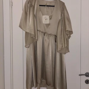 Champagnefärgad omlottklänning från Aéryne - Elegant champagnefärgad klänning från Aéryne med v-ringning och omlottdetalj. Klänningen har vida, fladdrande ärmar och ett mjukt, glansigt material som ger en lyxig känsla. Perfekt för dig som vill sticka ut med en stilren look. Prislapp kvar