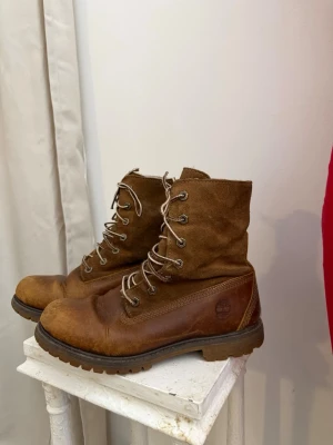 Timberland bruna kängor - Säljer ett par bruna kängor från Timberland i skinn och mocka. De har grov sula, snörning med metallhål och klassisk logga på sidan. Perfekta för dig som gillar en robust och stilren look.