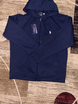 Marinblå hoodie från Polo Ralph Lauren - Snygg marinblå hoodie från Polo Ralph Lauren med dragkedja framtill och klassisk vit logga på bröstet. Hoodien har huva med dragsko och två fickor framtill. Tillverkad i mjukt bomullsmaterial som känns skönt mot huden. Perfekt för en avslappnad och stilren look.