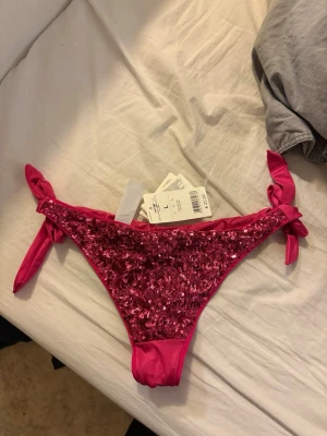 Calzedonia Rosa bikiniunderdel med paljetter - Snygg rosa bikiniunderdel från calzedonia med glittriga paljetter framtill och knyt i sidorna. Perfekt för dig som vill sticka ut på stranden eller vid poolen. Materialet är mjukt och stretchigt för en bekväm passform. Underdel L som är liten i storleken, överdelen 80B/75c/70d. Köpt för 550 euro, säljer för 450kr.