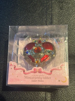 Sailor Moon hjärtformad smyckesask - Säljer en hjärtformad smyckesask från Sailor Moon-serien, tillverkad av Bandai. Asken är röd med guldfärgade detaljer och färgglada stenar, och kommer i originalförpackning med texten 'Miniaturely tablet'. Perfekt för fans av anime och samlarsaker.