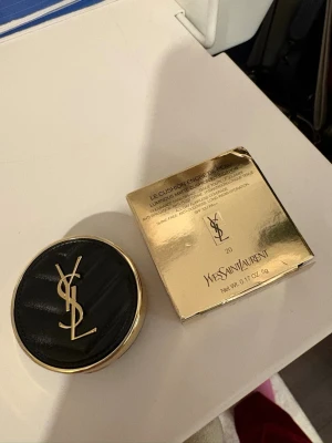 YSL Le Cushion Encre De Peau Foundation - YSL Le Cushion Encre De Peau Foundation i färg 20. Lyxig foundation i kuddförpackning med svart och guld design samt YSL-logga. Ger en naturlig och strålande finish. Kommer i en elegant rund dosa och med originalförpackning.