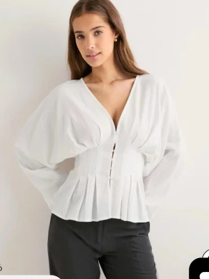 Vit v-ringad blus med knappar - Snygg vit blus med v-ringning och dekorativa knappar framtill. Blusen har långa, puffiga ärmar och markerad midja med plisserade detaljer. Perfekt för dig som vill ha en stilren och trendig look. Storlek 32 från Nelly, helt slutsåld. Säljer då den tyvärr var för liten. Dvs endast testad med prislappen kvar!!