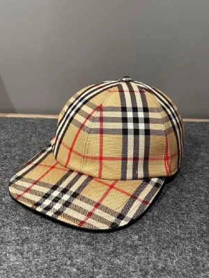 Beige rutig keps från Burberry - Säljer en klassisk keps från Burberry i beige med det ikoniska rutmönstret i svart, vitt och rött. Kepsen har böjd skärm och är tillverkad i ett slitstarkt tyg som ger en exklusiv känsla. Perfekt accessoar för dig som gillar streetstyle och designerplagg.