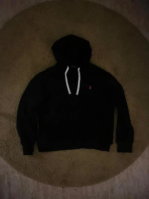 Svart hoodie från Polo Ralph Lauren - Svart hoodie från Polo Ralph Lauren med vit dragsko och liten röd logga broderad på bröstet. Klassisk passform med huva och långa ärmar. Perfekt för en chill och stilren look. Inte använd så mycket. Storlek S men passar bättre för XS.