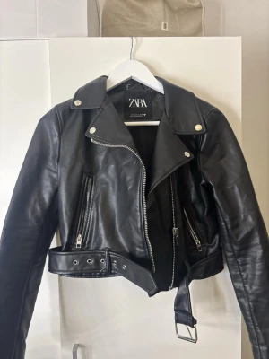 Svart skinnjacka från Zara - Snygg svart skinnjacka från Zara med klassisk bikerstil, silvriga dragkedjor och detaljer samt bälte i midjan. Jackan har bred krage och två snedställda fickor med dragkedja. Perfekt för en cool och trendig look. Fint skick. 