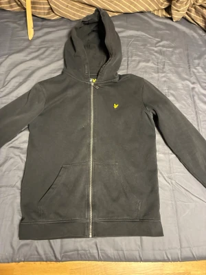 Svart hoodie från Lyle & Scott - Säljer en svart hoodie med dragkedja från Lyle & Scott. Klassisk design med huva, två fickor framtill och det ikoniska gula fågel-loggan broderad på bröstet. Perfekt för en chill och stilren look. Materialet är mjuk bomullsmix som känns skön mot huden.