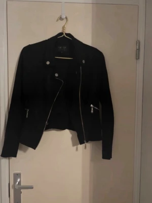 Svart skinnjacka med dragkedjor - Snygg svart skinnjacka från women colletion med asymmetrisk dragkedja, silverfärgade knappar och detaljer. Jackan har två fickor med dragkedja framtill och en kort, figurnära passform som ger en cool look.