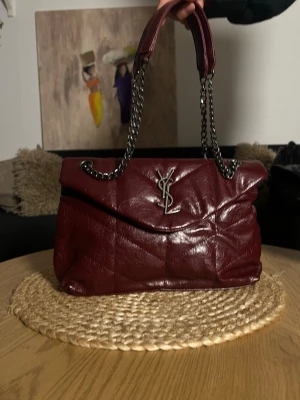 Vinröd YSL handväska i controläder - Säljer en vinröd handväska från Saint Laurent i quiltat läder med silverfärgad kedjerem och ikonisk YSL-logga framtill. Väskan har en mjuk, puffig form och stängs med lock. Perfekt för dig som vill ha en lyxig och trendig accessoar.