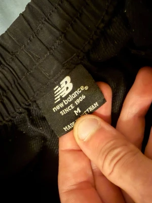 Svarta shorts från New Balance - Snygga svarta shorts från New Balance med elastisk midja och stort logotryck längs benet. Perfekta för träning eller chill, tillverkade i ett mjukt och lätt material som känns skönt mot huden.