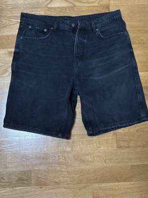 Svarta jeansshorts i loose fit - Säljer ett par svarta jeansshorts med klassisk femficksdesign och hällor för bälte. Shortsen har en loose fit och är tillverkade i denim, perfekta för en avslappnad stil. Passar dig som gillar enkel och tidlös look.