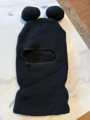Svart balaklava med öron - Säljer en svart stickad balaklava med två runda öron på toppen. Mössan har ett avlångt ansiktsöppning och är gjord i mjukt, ribbstickat material. Perfekt för dig som vill ha en lekfull och unik look under kalla dagar.