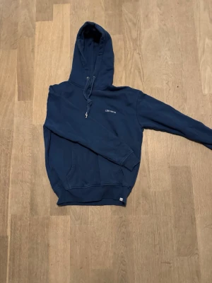 Mörkblå hoodie från Les Deux - Snygg mörkblå hoodie från Les Deux med liten vit logga på bröstet. Klassisk modell med huva och känguruficka framtill. Perfekt för en chill och avslappnad stil. Mjuk bomullskänsla och ribbade muddar vid ärmslut och nederkant.