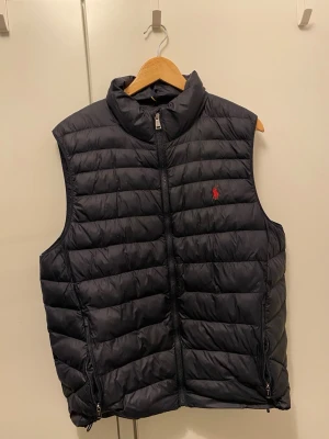 Svart dunväst från Polo Ralph Lauren - Snygg svart dunväst från Polo Ralph Lauren med quiltad design och hög krage. Västen har dragkedja framtill, två fickor med dragkedja och den klassiska röda loggan broderad på bröstet. Perfekt för lager-på-lager och riktigt stilren.