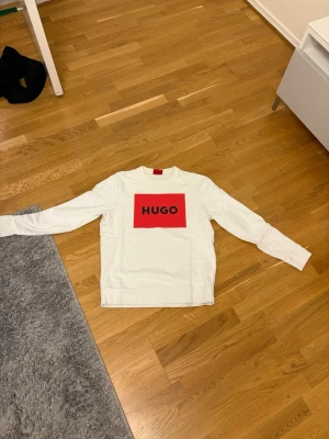 Vit sweatshirt från Hugo med logga - Säljer en vit sweatshirt från Hugo med stor röd logga på bröstet. Tröjan har rund halsringning och långa ärmar. Perfekt för dig som gillar enkel men snygg streetwear med tydlig branding.