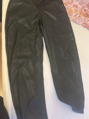 Svarta fuskskinnjeans med raka ben - Säljer ett par svarta jeans i fuskskinn med raka ben och klassisk femficksdesign. Byxorna har hög midja och stängs med dragkedja och knapp. Perfekta för dig som vill ha en edgy look och gillar trendiga material.