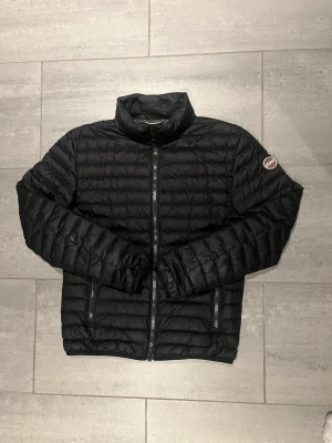 Svart pufferjacka från Colmar - Snygg svart pufferjacka från Colmar med hög krage och dragkedja framtill. Jackan har två sidofickor med dragkedja och Colmar-logga på ena ärmen. Perfekt för kalla dagar och riktigt skön att bära.storlek 46