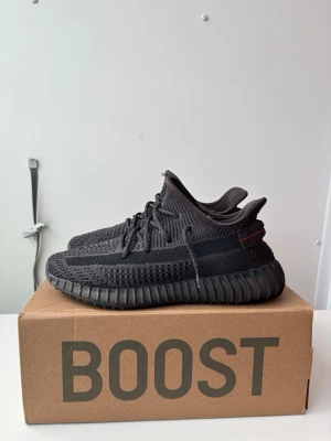 Yeezy Boost 350 V2 Black sneakers - Svarta Yeezy Boost 350 V2 från Adidas med stickad ovandel i mesh, svart sula och diskreta röda detaljer på hälen. Skorna har en chunky siluett, snörning och den klassiska BOOST-sulan för extra komfort. Perfekta för dig som gillar streetwear och vill sticka ut.