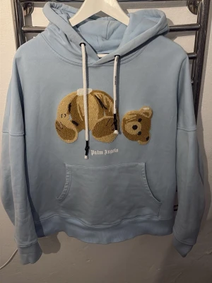 Ljusblå hoodie från Palm Angels - Ljusblå hoodie från Palm Angels med stor broderad nallebjörn på bröstet och vit logotyp under. Hoodien har huva med vita dragsnören och en stor magficka framtill. Tillverkad i mjukt bomullsmaterial, perfekt för en avslappnad streetwear-look.