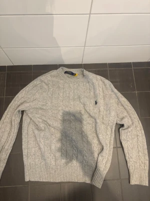 Grå kabelstickad tröja Polo Ralph Lauren L - Kabelstickad grå tröja från Polo Ralph Lauren med rund halsringning och broderad logga på bröstet. Tröjan har ribbade muddar vid ärmslut och nederkant. Perfekt för lager-på-lager och klassisk stil.