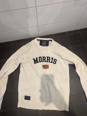 Vit tröja från Morris med tryck M - Vit långärmad tröja från Morris med svart textlogga och brittisk flagga broderad på bröstet. Tröjan har rund halsringning och ribbade muddar vid ärmslut och nederkant. Perfekt för en stilren och klassisk look.