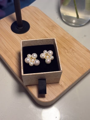 Ioaku örhängen  - Säljer ett par örhängen i form av blommor med vita pärlor och små guldfärgade detaljer i mitten. Örhängena har en elegant och söt design som verkligen sticker ut. Perfekta för dig som gillar klassiska accessoarer med en twist.