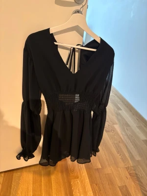 Svart klänning med v-ringning och knytband - Snygg svart klänning med v-ringning och knytband i nacken. Klänningen har långa, lätt transparenta ärmar med resår vid handlederna och smock i midjan för en markerad siluett. Tillverkad i ett tunt, flowy material som ger en luftig känsla.