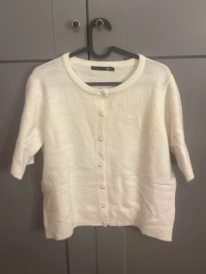 Vit kofta med pärlknappar från H&M - Mysig vit kofta från H&M med rund halsringning och pärlknappar framtill. Koftan har halvlånga ärmar och är stickad i ett mjukt material. Perfekt att slänga över en topp för en clean och stilren look.
