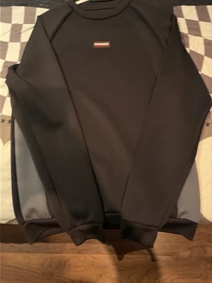 Svart sweatshirt från McKenzie  - Säljer en svart sweatshirt från McKenzie med gråa paneler på sidorna. Tröjan har rund halsringning och en liten logotyp på bröstet. Perfekt för en avslappnad och stilren look.