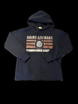 Mörkblå Saint Laurent hoodie - Snygg mörkblå hoodie från Saint Laurent med tryck framtill i vitt och rött samt logga i mitten. Klassisk känguruficka och justerbar huva med snören. Tillverkad i mjukt bomullsmaterial, perfekt för en avslappnad och trendig look. Tröja såklart äkta, har taggar kvar! Perfekt för dig som är ca 164 cm lång, säljer då det är för litet