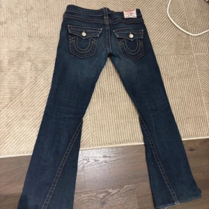 True Religion mörkblå bootcut jeans - Mörkblå jeans från True Religion med bootcut passform och kontrastsömmar. Snygga fickdetaljer bak med vita knappar och klassisk logopatch i linningen. Tillverkade i denim med lätt slitning för en cool look.
