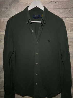 Mörkgrön skjorta från Polo Ralph Lauren - Stilren mörkgrön skjorta från Polo Ralph Lauren med klassisk krage och knappar framtill. Skjortan har lång ärm och den ikoniska broderade loggan på bröstet. Perfekt för dig som gillar en clean och tidlös look.